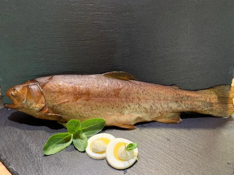 Kräftig geräucherter ganzer Fisch mit dem Kopf nach links, davor garnierte Ei-Hälften und frischer Oregano.